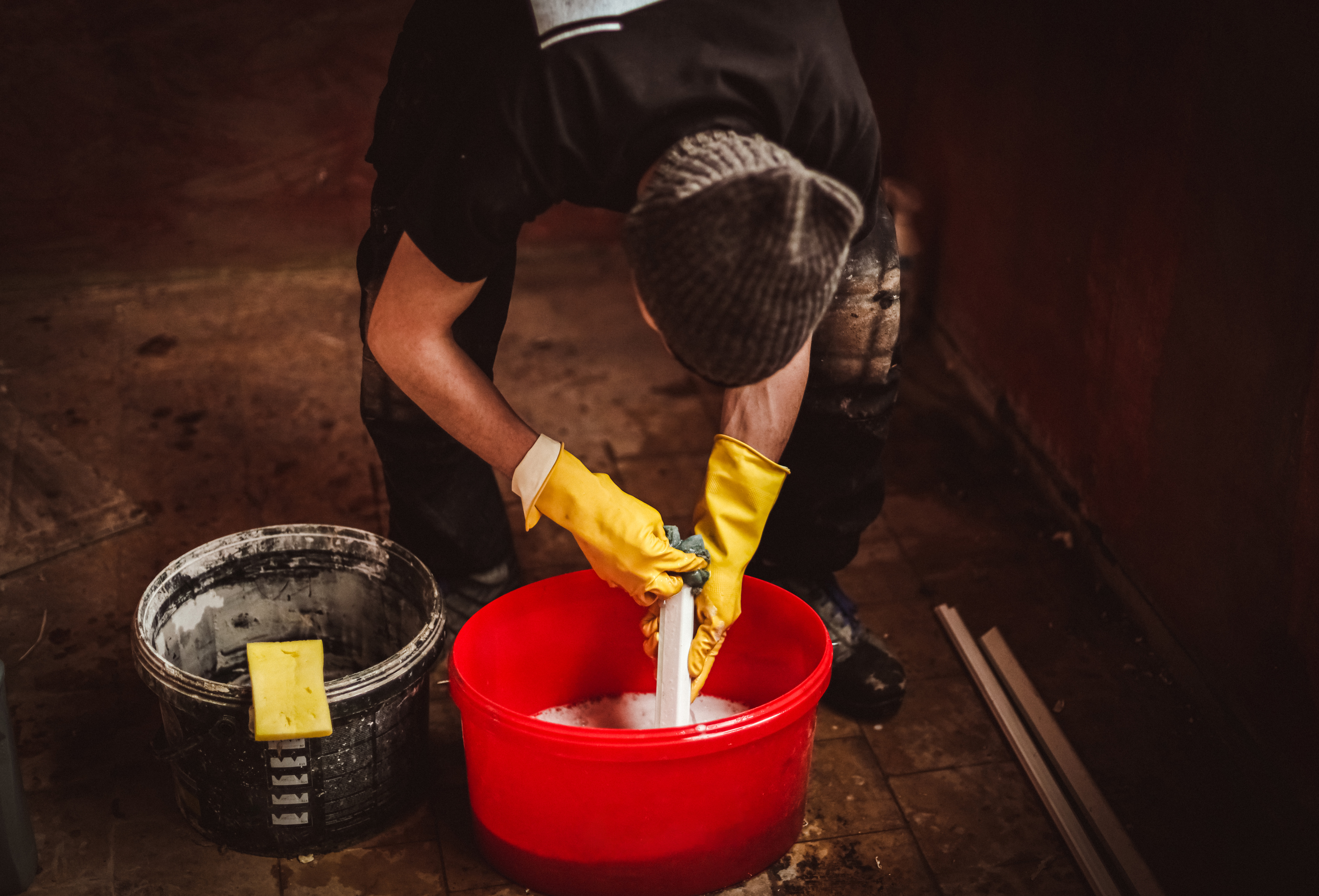 One young Caucasian man in yellow gloves, stands bent over and washes with detergent using a sponge part of a plastic window frame in a red bucket in a dark house, top side close-up view with selective focus. Concept of home renovation and washing window frames.